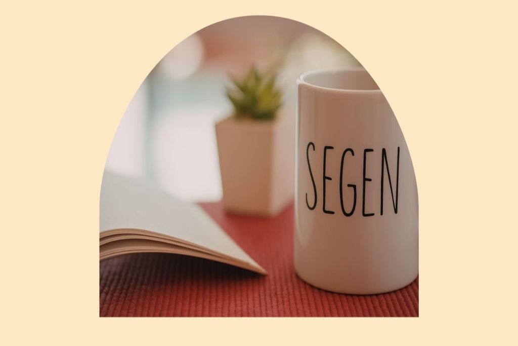 Ein aufgeschlagenes Buch, eine Tasse mit dem Schriftzug Segen und eine kleinen Pflanze im Hintergrund.